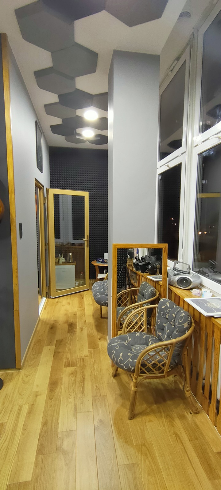 Wnętrze studia nagrań z panelami akustycznymi na ścianie, drewnianą podłogą i wiklinowymi fotelami z szarymi poduszkami w kwiaty, widok na okno i sprzęt nagraniowy.