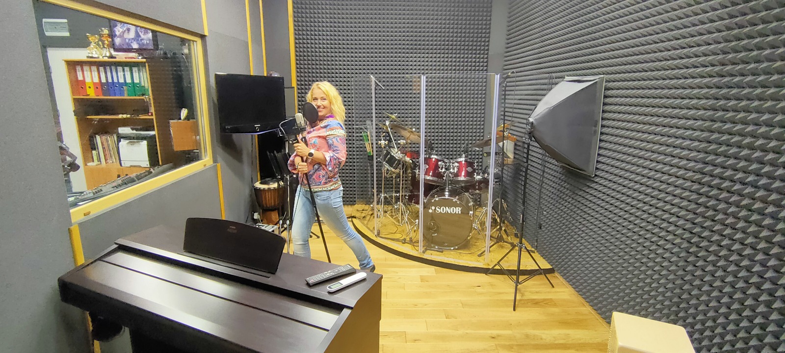 Kobieta z mikrofonem w ręku, w profesjonalnym studio nagrań z pianinem, perkusją za plecami i panelami akustycznymi na ścianach.