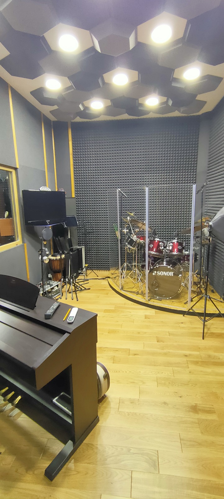 Wnętrze studia nagrań z pianinem cyfrowym, perkusją Sonor oddzieloną szybą akustyczną, monitorami studyjnymi i panelami akustycznymi na ścianach i suficie.
