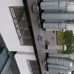 Usługi kominiarsko-Gazowe Aleksander Ostrowski - Szereg metalowych wylotów wentylacyjnych na płaskim dachu budynku, zabezpieczonych gumowymi kołnierzami, widoczne kratki wentylacyjne w ścianie.