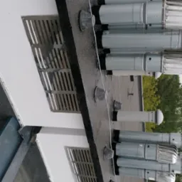 Szereg metalowych wylot&oacute;w wentylacyjnych na płaskim dachu budynku, zabezpieczonych gumowymi kołnierzami, widoczne kratki wentylacyjne w ścianie.