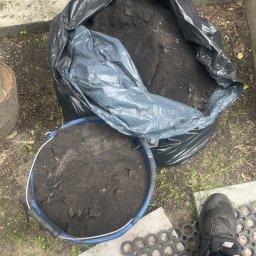 Usługi kominiarsko-Gazowe Aleksander Ostrowski - Worek i wiadro z czarną ziemią w Żyrardowie, cegły w tle. Przygotowanie do prac ziemnych, widoczne fragmenty butów i maty gumowej.