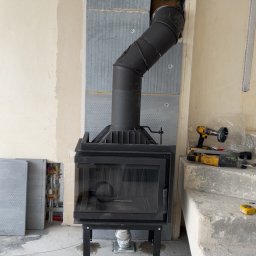 Usługi kominiarsko-Gazowe Aleksander Ostrowski - Czarny kominek z rurą dymową w trakcie montażu, widoczne narzędzia i materiały budowlane, surowy stan wykończenia wnętrza.