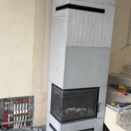 Usługi kominiarsko-Gazowe Aleksander Ostrowski - Surowy kominek z szarej płyty, w trakcie obudowy, z widocznymi elementami instalacji grzewczej w tle, na tle surowej ściany i schodów.