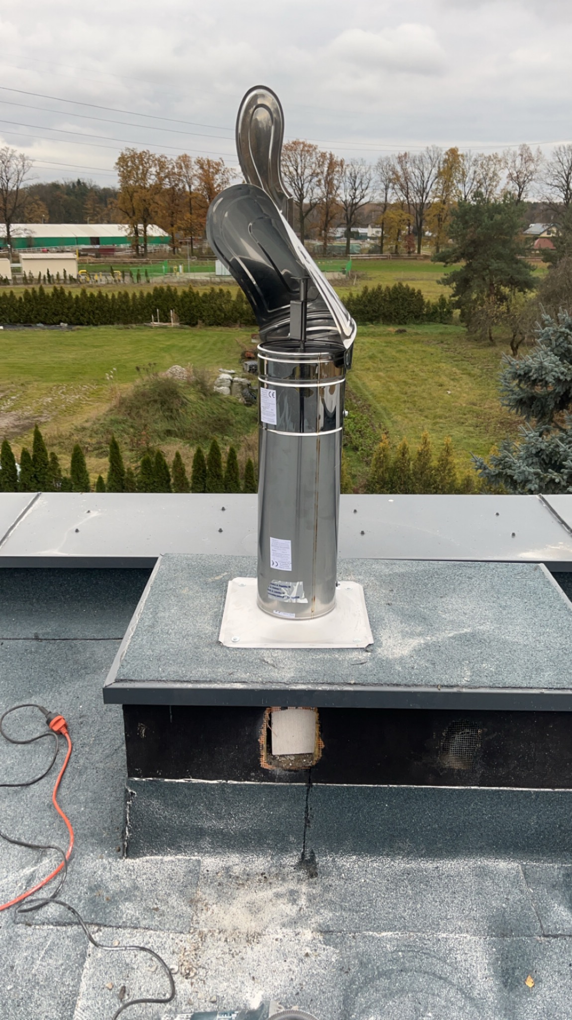 Błyszczący, metalowy wkład kominowy z daszkiem na dachu, widok z góry na tle zieleni i pochmurnego nieba. Widoczne ślady prac montażowych na dachu, w pobliżu podłączenie elektryczne.