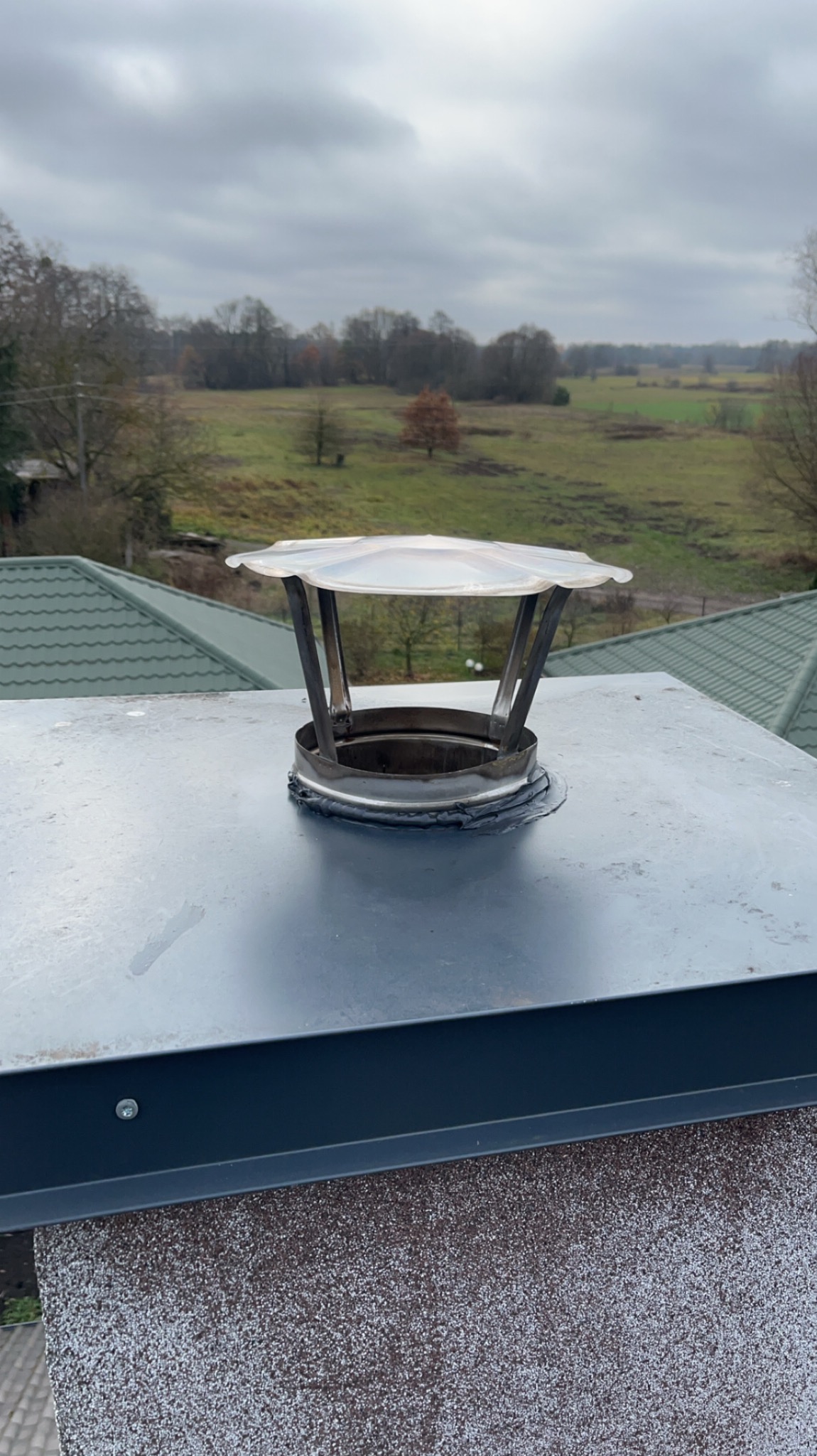 Metalowy komin z daszkiem ochronnym na dachu z widokiem na polną scenerię w pochmurny dzień.