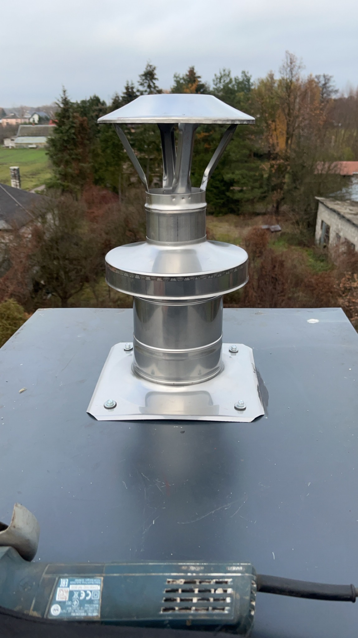 Nowy, metalowy komin z daszkiem zamontowany na ciemnym dachu, widoczny z bliska, z fragmentem szlifierki kątowej w dolnym rogu. W tle drzewa i zabudowania.