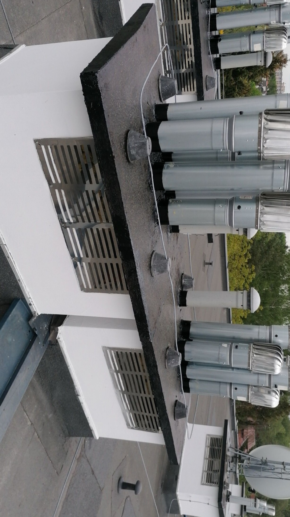 Szereg metalowych wylotów wentylacyjnych na płaskim dachu budynku, zabezpieczonych gumowymi kołnierzami, widoczne kratki wentylacyjne w ścianie.