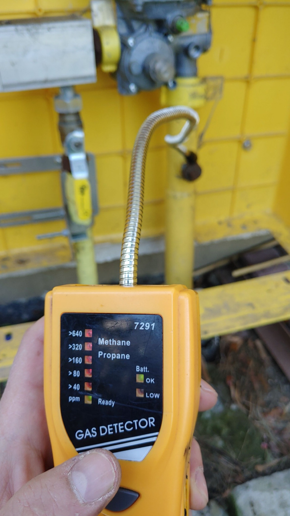 Żółty detektor gazu z elastyczną sondą, wskazujący obecność metanu i propanu, na tle żółtej instalacji gazowej.
