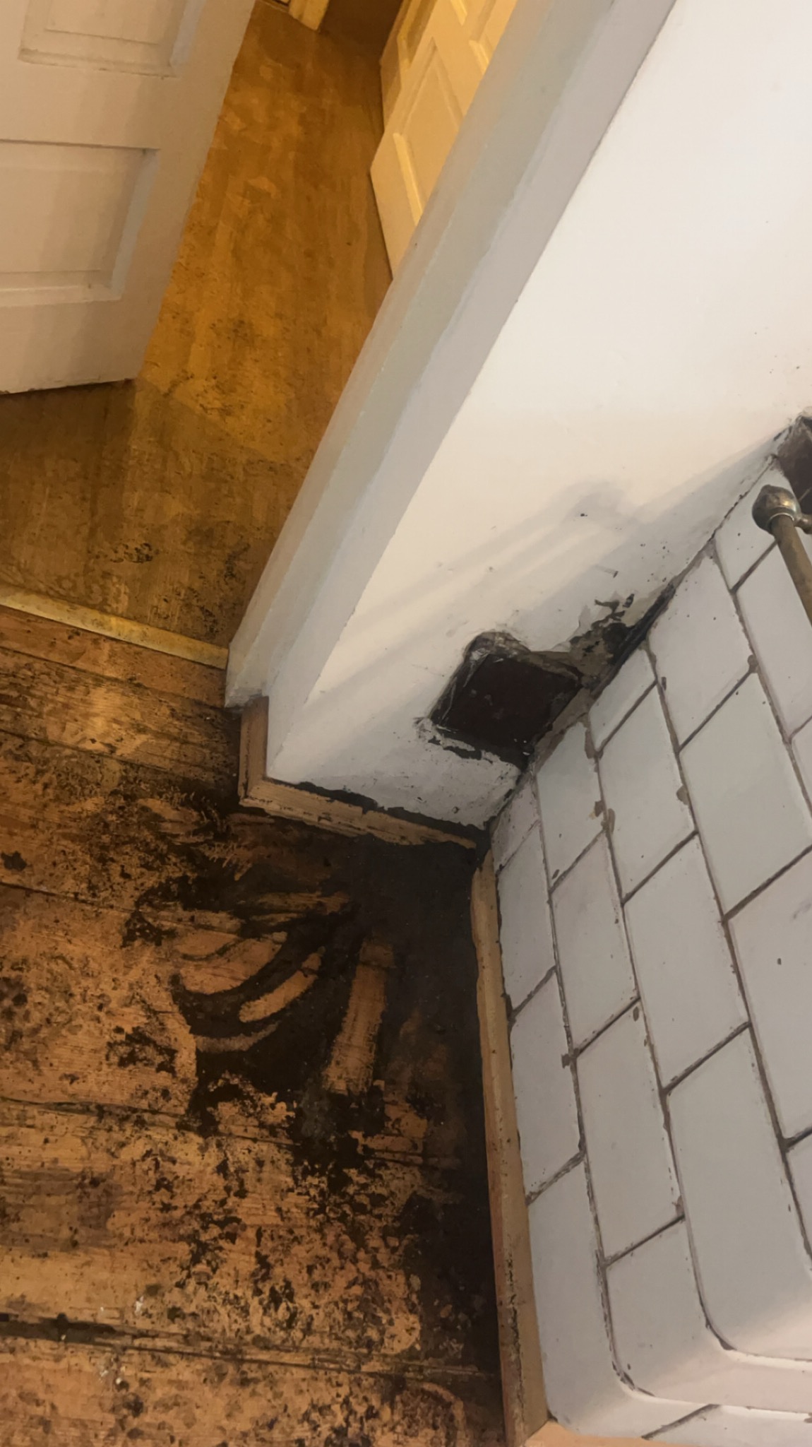 Narożnik pomieszczenia z drewnianą podłogą i fragmentem białych płytek. Widoczny otwór w ścianie z pozostałościami po instalacji, prawdopodobnie wentylacyjnej.