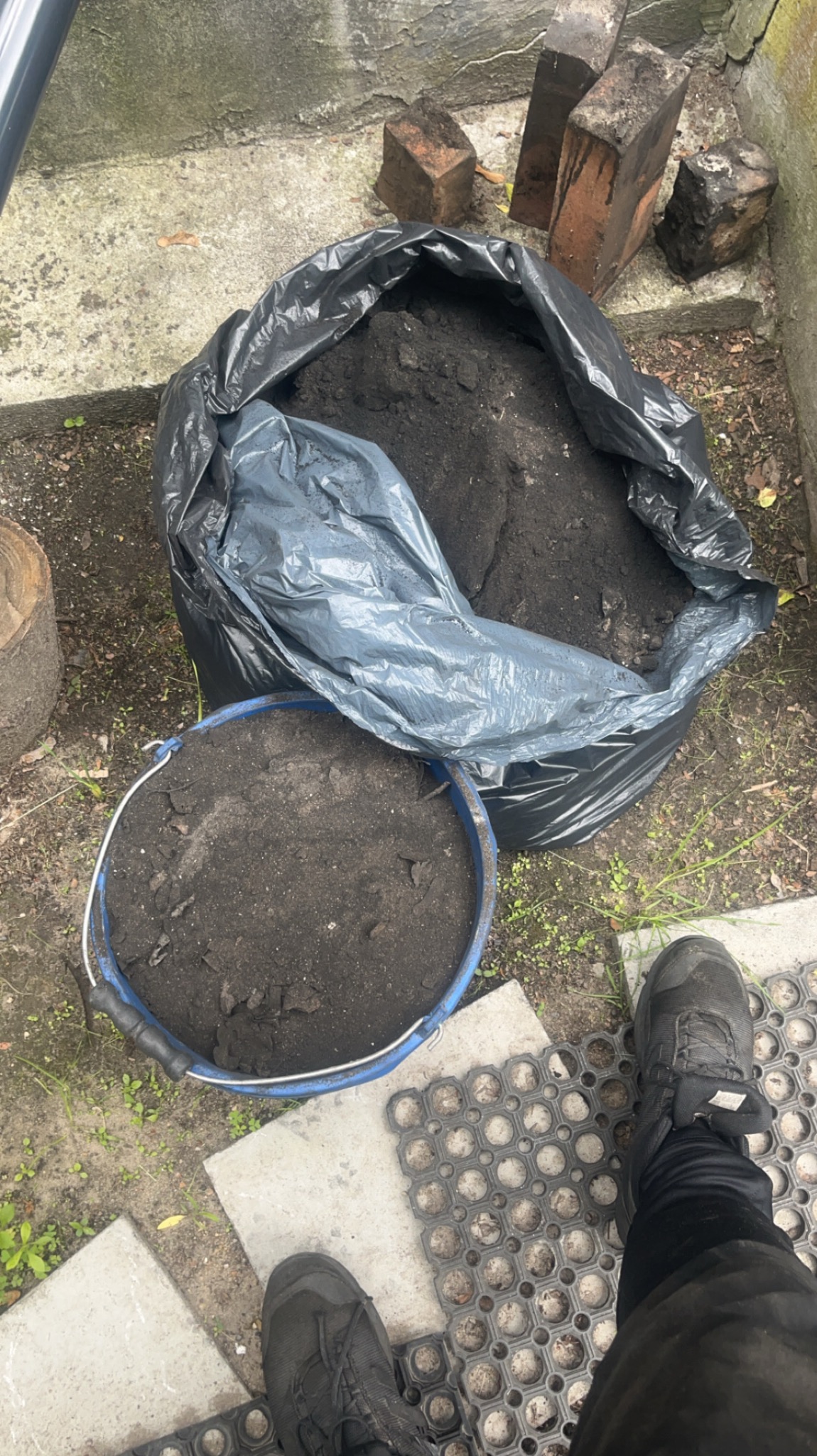 Worek i wiadro z czarną ziemią w Żyrardowie, cegły w tle. Przygotowanie do prac ziemnych, widoczne fragmenty butów i maty gumowej.