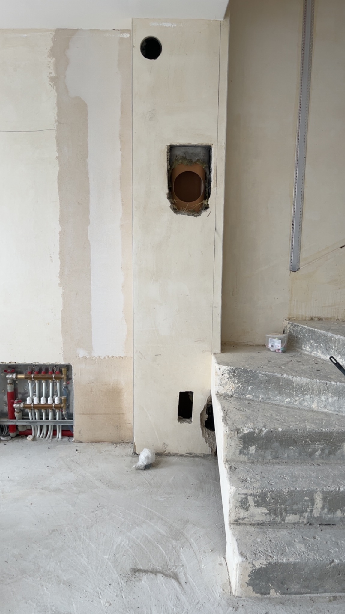 Surowy stan wnętrza: instalacja CO z rozdzielaczem, otwory w ścianie pod wentylację i kominek, betonowe schody. Widoczne ślady po pracach budowlanych.
