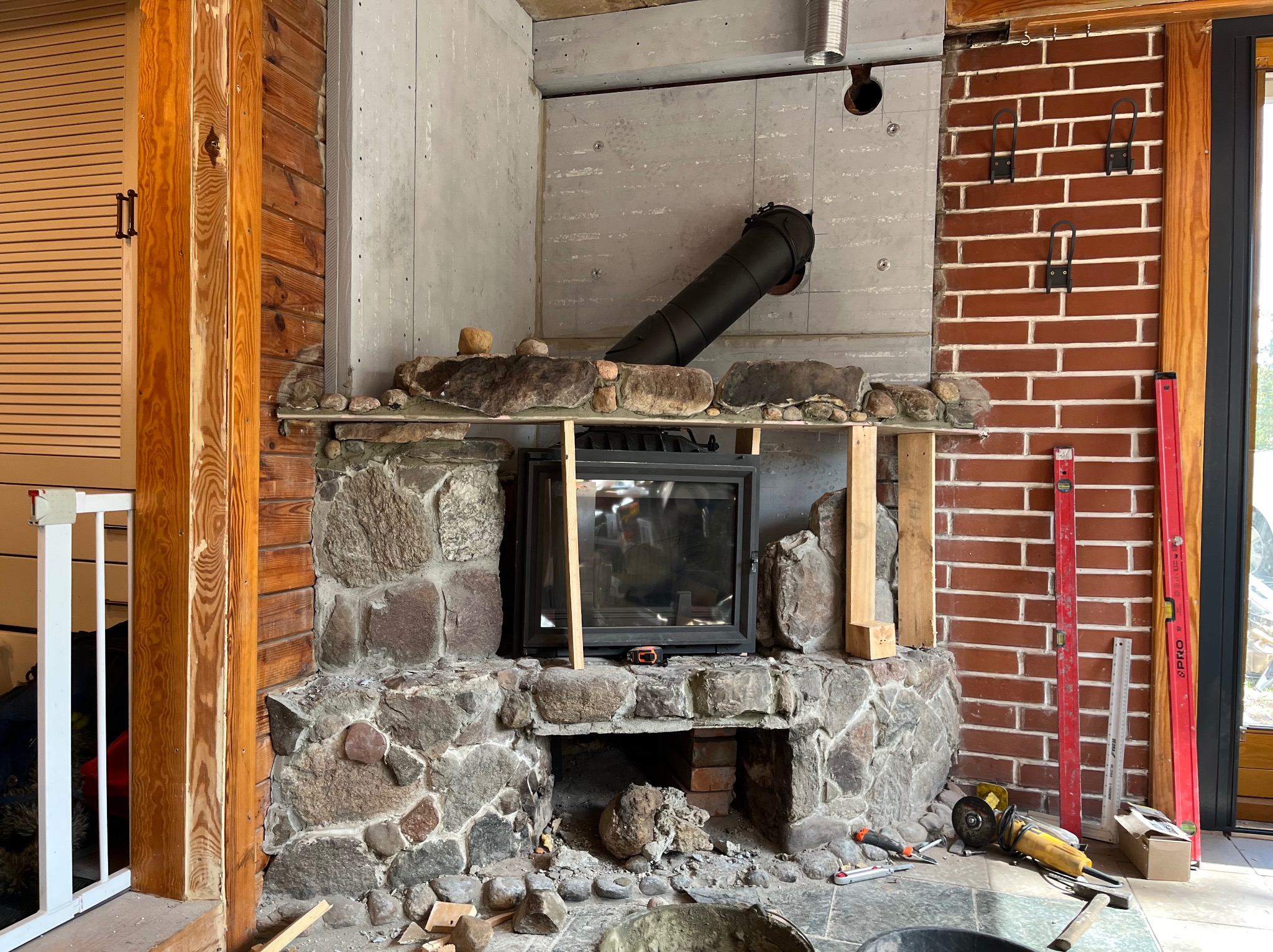 Kamienny kominek w trakcie budowy z widocznym wkładem kominkowym, rurą odprowadzającą dym i narzędziami. Surowe wykończenie, cegła i drewno w tle.