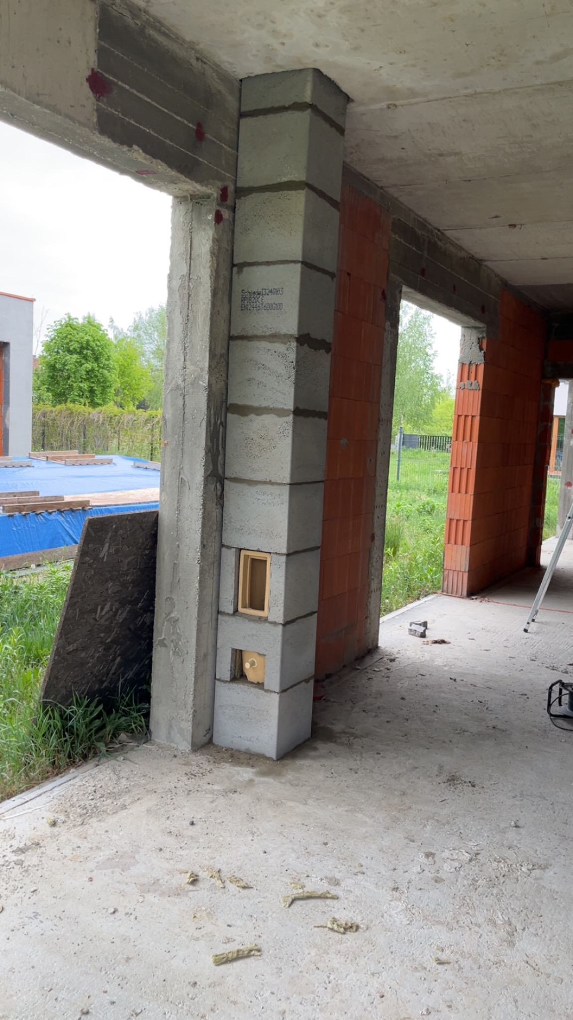 Wznoszona konstrukcja komina z pustaków szamotowych z widocznymi otworami rewizyjnymi, na tle surowych ścian z czerwonej cegły i betonowych filarów w stanie deweloperskim.