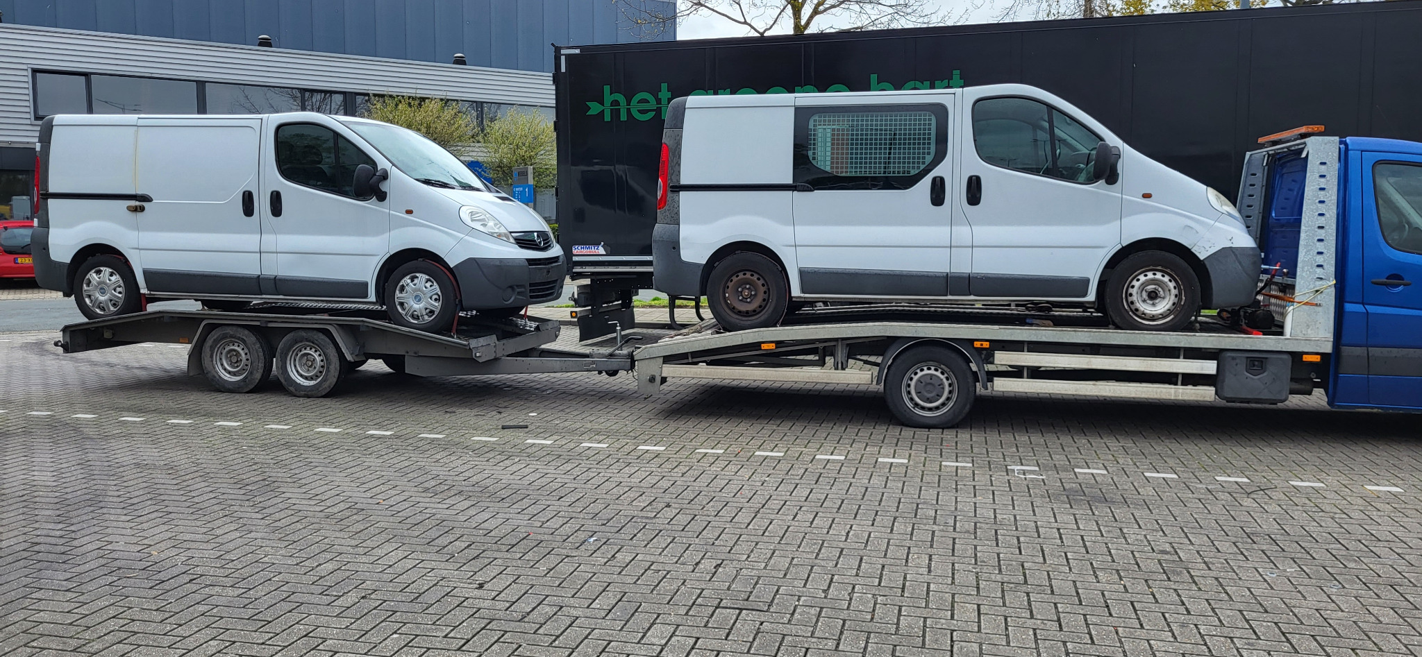 Dwa białe busy marki Opel Vivaro przewożone na lawetach, jedna laweta ciągnięta przez niebieski samochód dostawczy, na tle szary budynek i czarna naczepa z zielonym napisem.