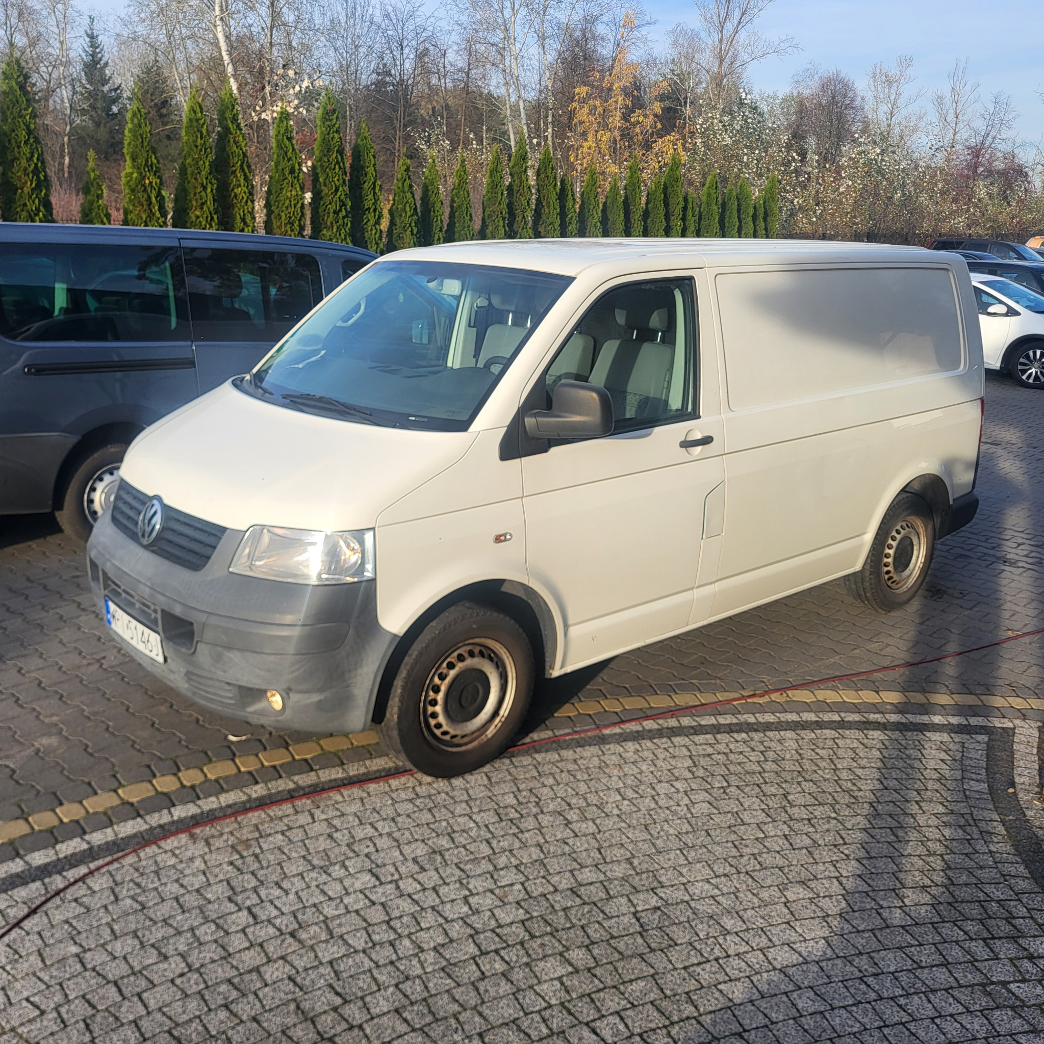 Biały samochód dostawczy Volkswagen Transporter T5 zaparkowany na brukowanym parkingu, widoczne inne pojazdy i drzewa w tle.