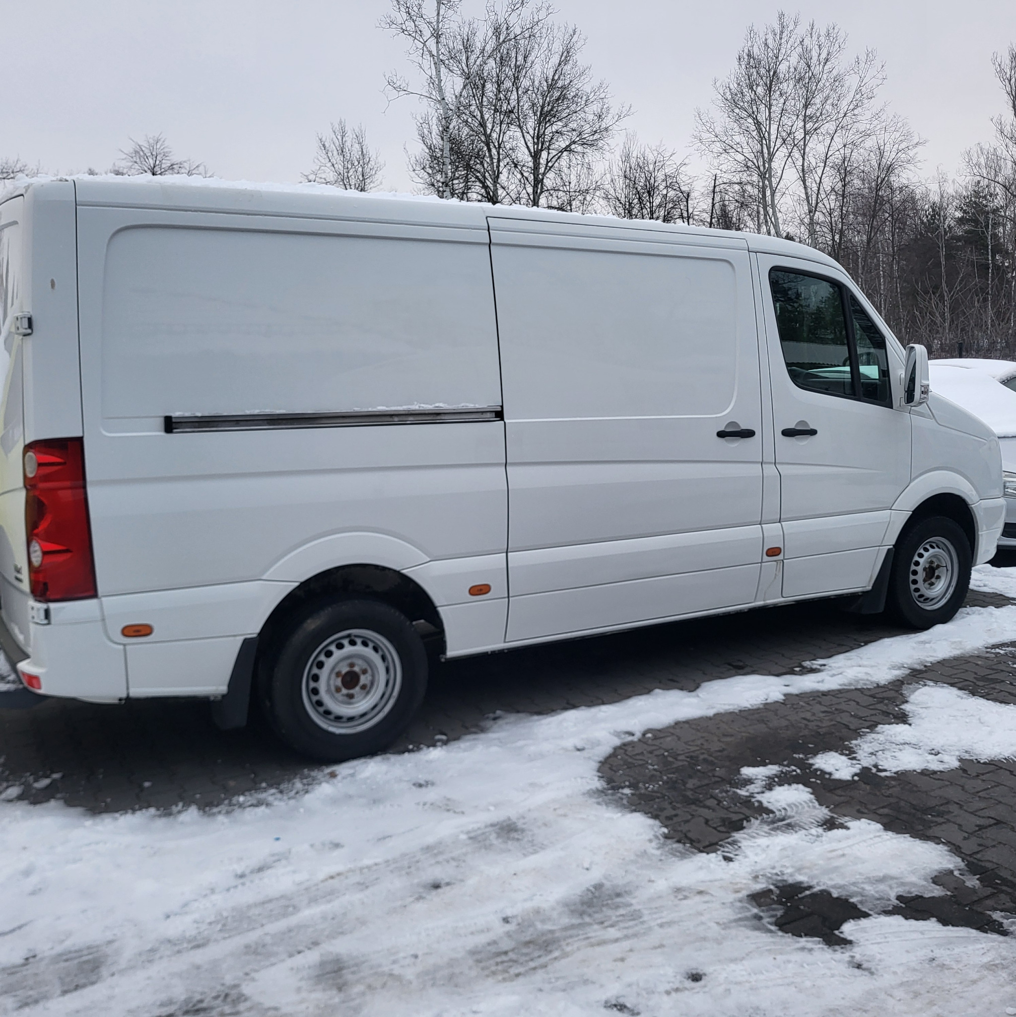 Biały samochód dostawczy zaparkowany na parkingu z kostki brukowej pokrytym śniegiem, widok z boku, w tle drzewa bez liści pod pochmurnym niebem.