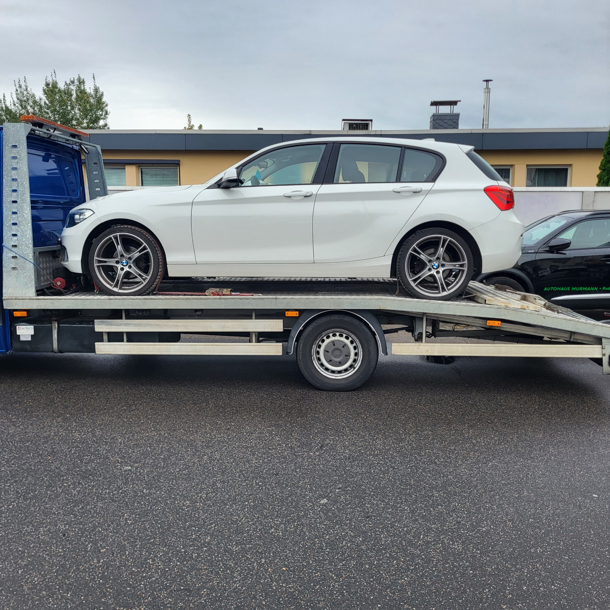 Biały samochód BMW serii 1 na niebieskiej lawecie, widok z boku, na mokrym asfalcie, w tle budynek i zaparkowany czarny samochód.