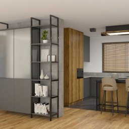 MAFF - Wnętrze mieszkania z otwartą kuchnią, drewnianą podłogą i schodami z czarnymi balustradami. Widoczna wyspa kuchenna z dwoma hokerami oraz regał z książkami i dekoracjami.