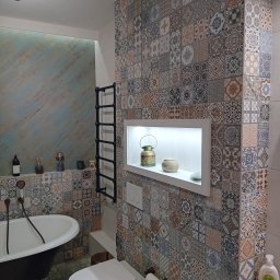 MAFF - Wnętrze łazienki z wolnostojącą wanną na lwich nóżkach, toaletą typu Geberit i ścianą wyłożoną patchworkowymi płytkami w odcieniach beżu, brązu i błękitu. Na ścianie czarny grzejnik drabinkowy...