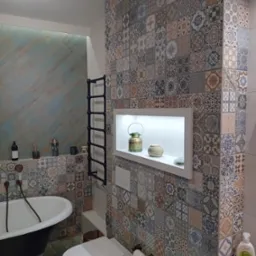 Wnętrze łazienki z wolnostojącą wanną na lwich nóżkach, toaletą typu Geberit i ścianą wyłożoną patchworkowymi płytkami w odcieniach beżu, brązu i błękitu. Na ścianie czarny grzejnik drabinkowy...