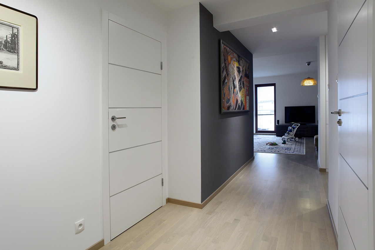 Nowoczesny korytarz z białymi drzwiami z poziomymi frezami, szarą ścianą z abstrakcyjnym obrazem i jasną podłogą z desek, widok w głąb pokoju z telewizorem i wózkiem dziecięcym.