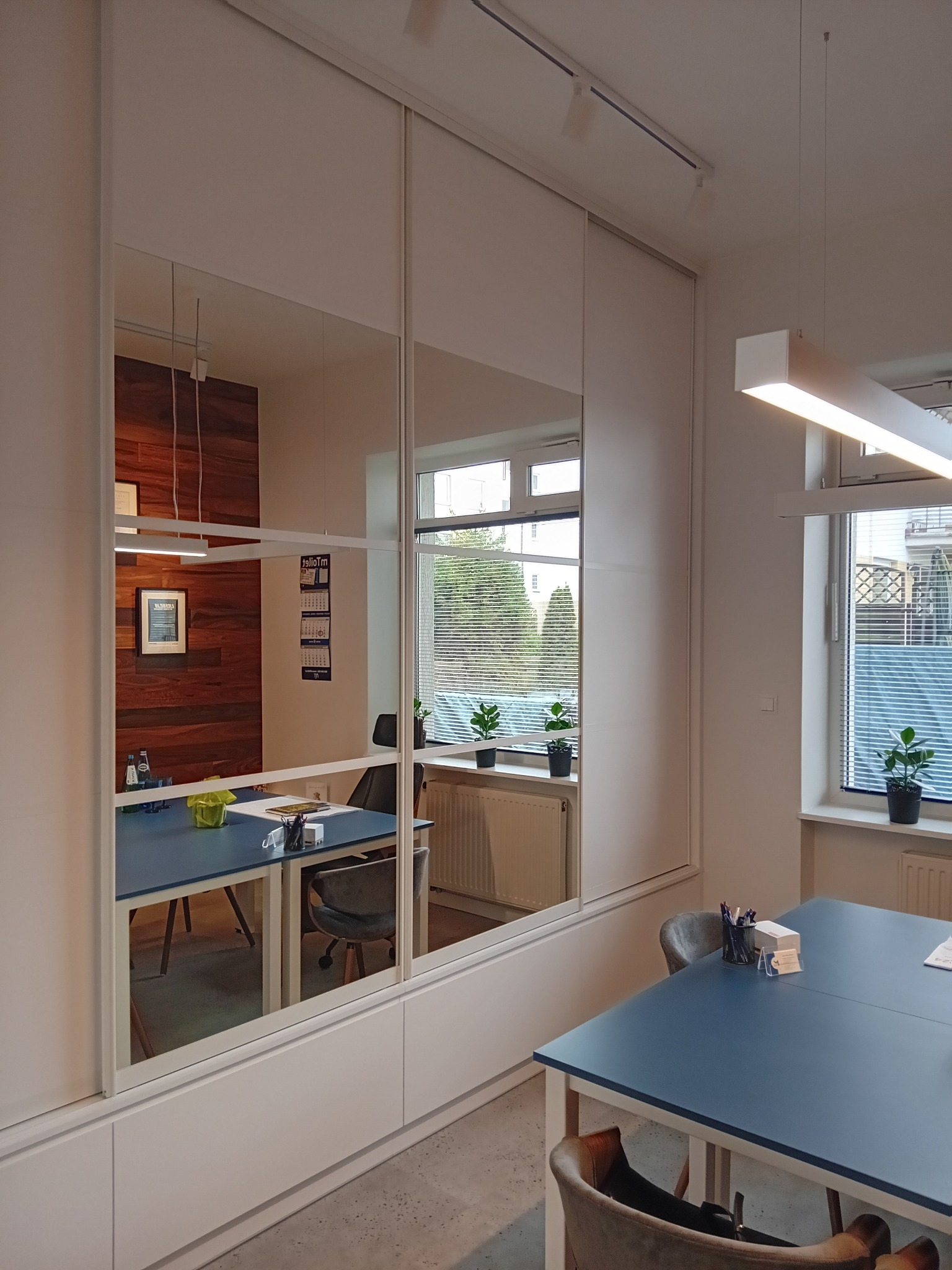 Nowoczesne biuro z dużym lustrem wbudowanym w szafę, odbijającym drewnianą ścianę i biurko z artykułami biurowymi.