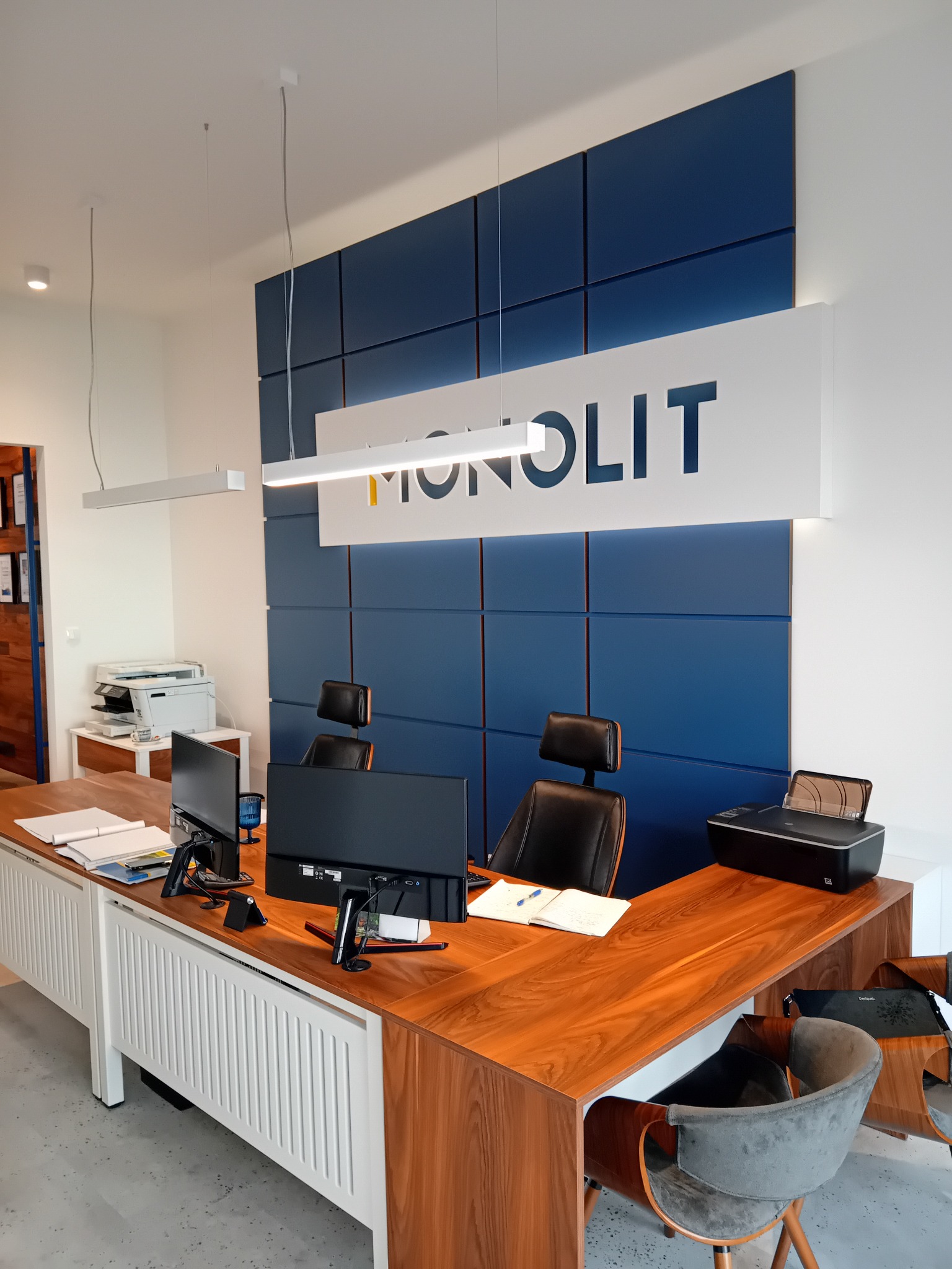 Nowoczesne biuro z drewnianym biurkiem, dwoma monitorami, drukarką i krzesłami na tle ściany z niebieskich paneli i podświetlanym logo firmy 'MONOLIT'.