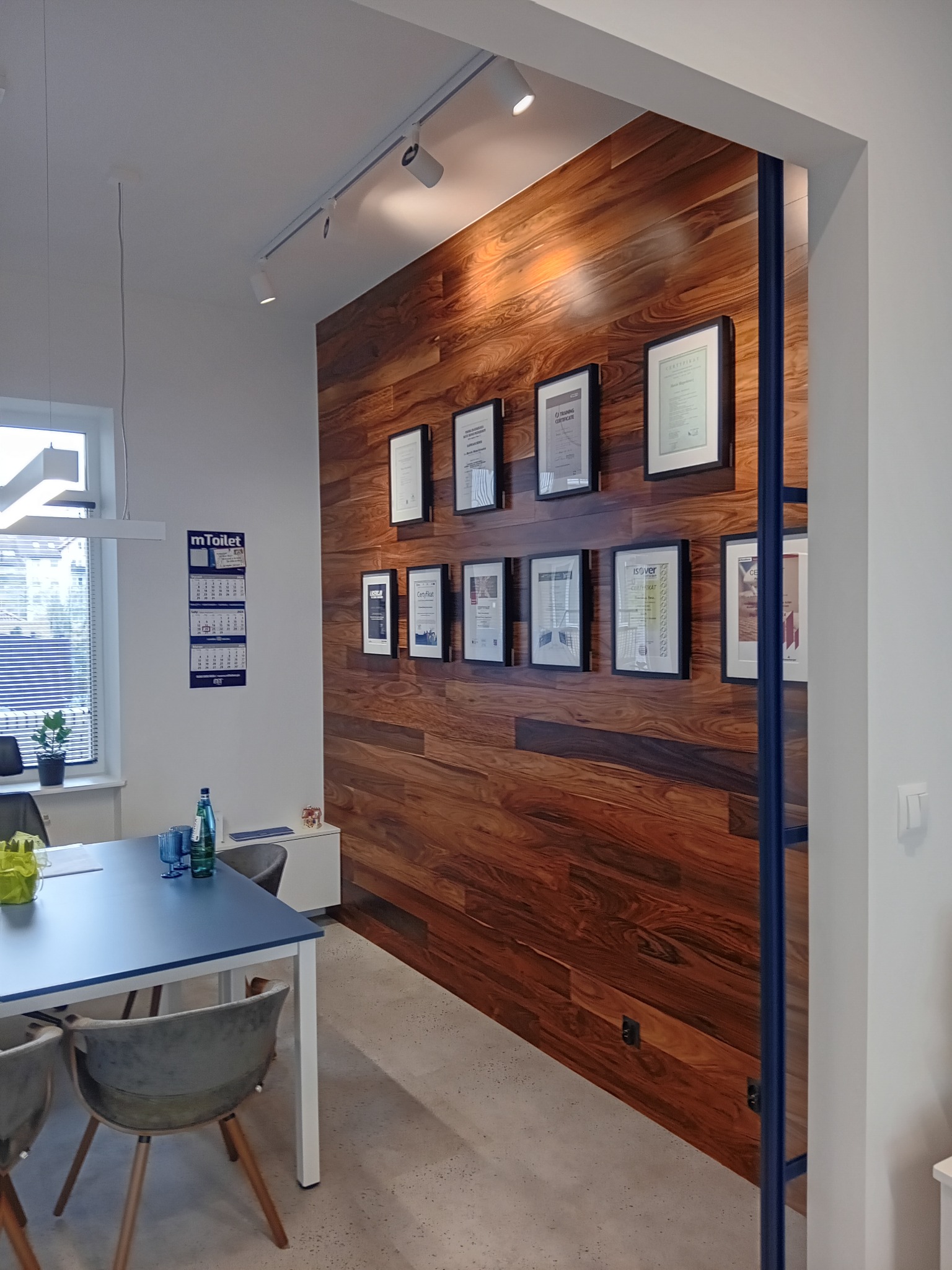 Biuro z drewnianą ścianą ozdobioną ramkami z certyfikatami, oświetleniem punktowym i niebieskim stołem konferencyjnym z szarymi krzesłami.