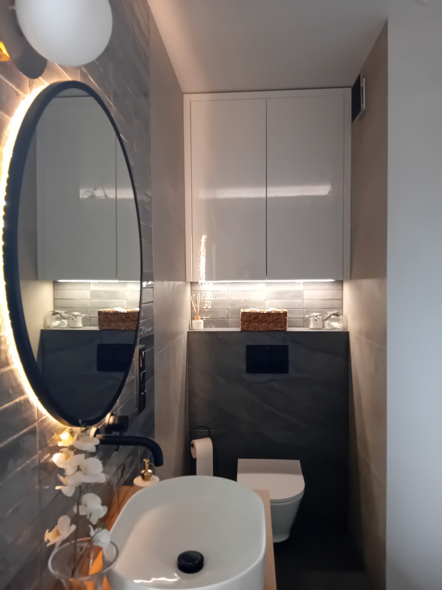 Nowoczesna łazienka z owalnym lustrem podświetlanym LED, umywalką nablatową, czarną armaturą i wiszącą szafką z oświetleniem punktowym.