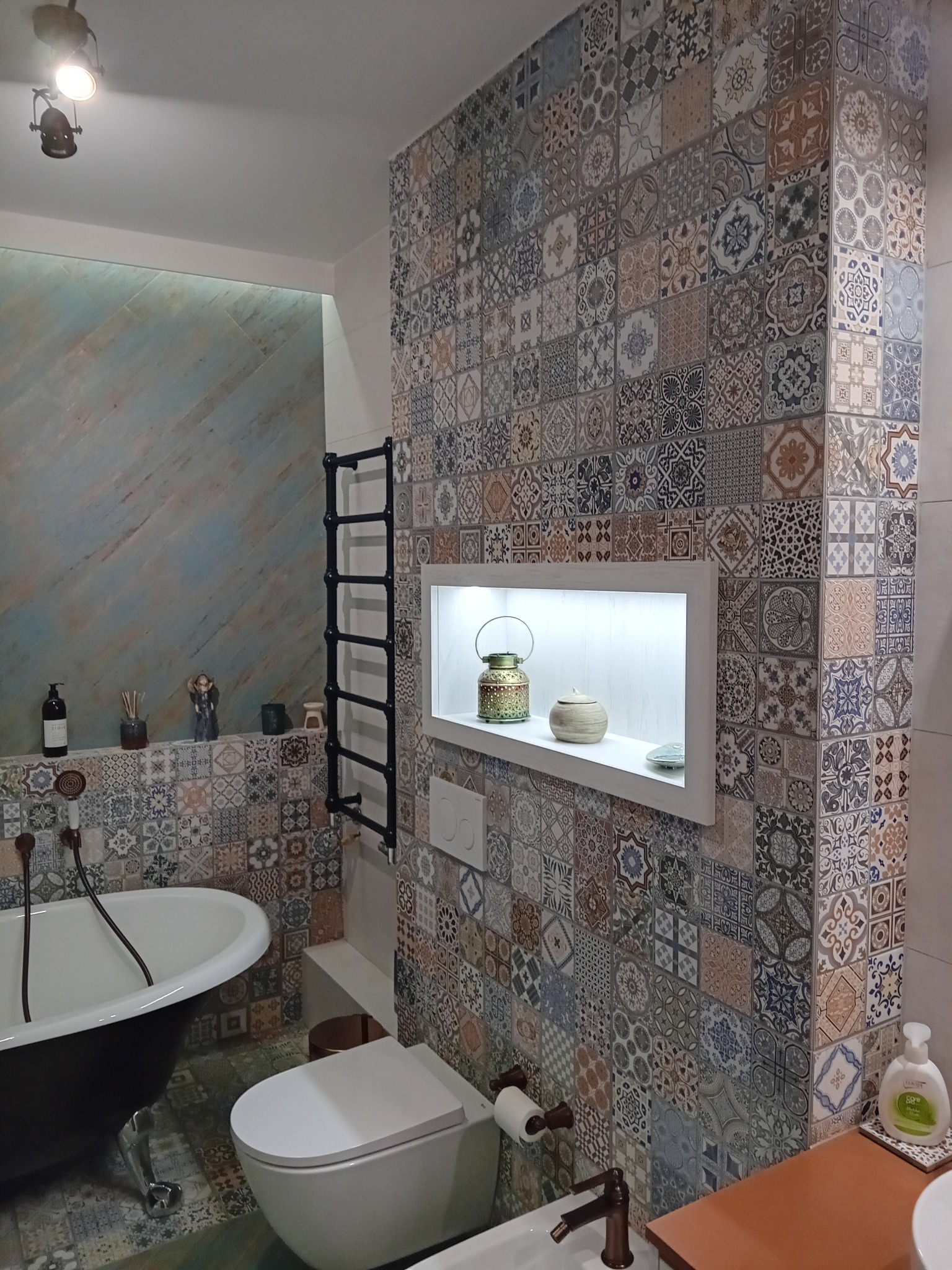 Wnętrze łazienki z wolnostojącą wanną na lwich nóżkach, toaletą typu Geberit i ścianą wyłożoną patchworkowymi płytkami w odcieniach beżu, brązu i błękitu. Na ścianie czarny grzejnik drabinkowy...