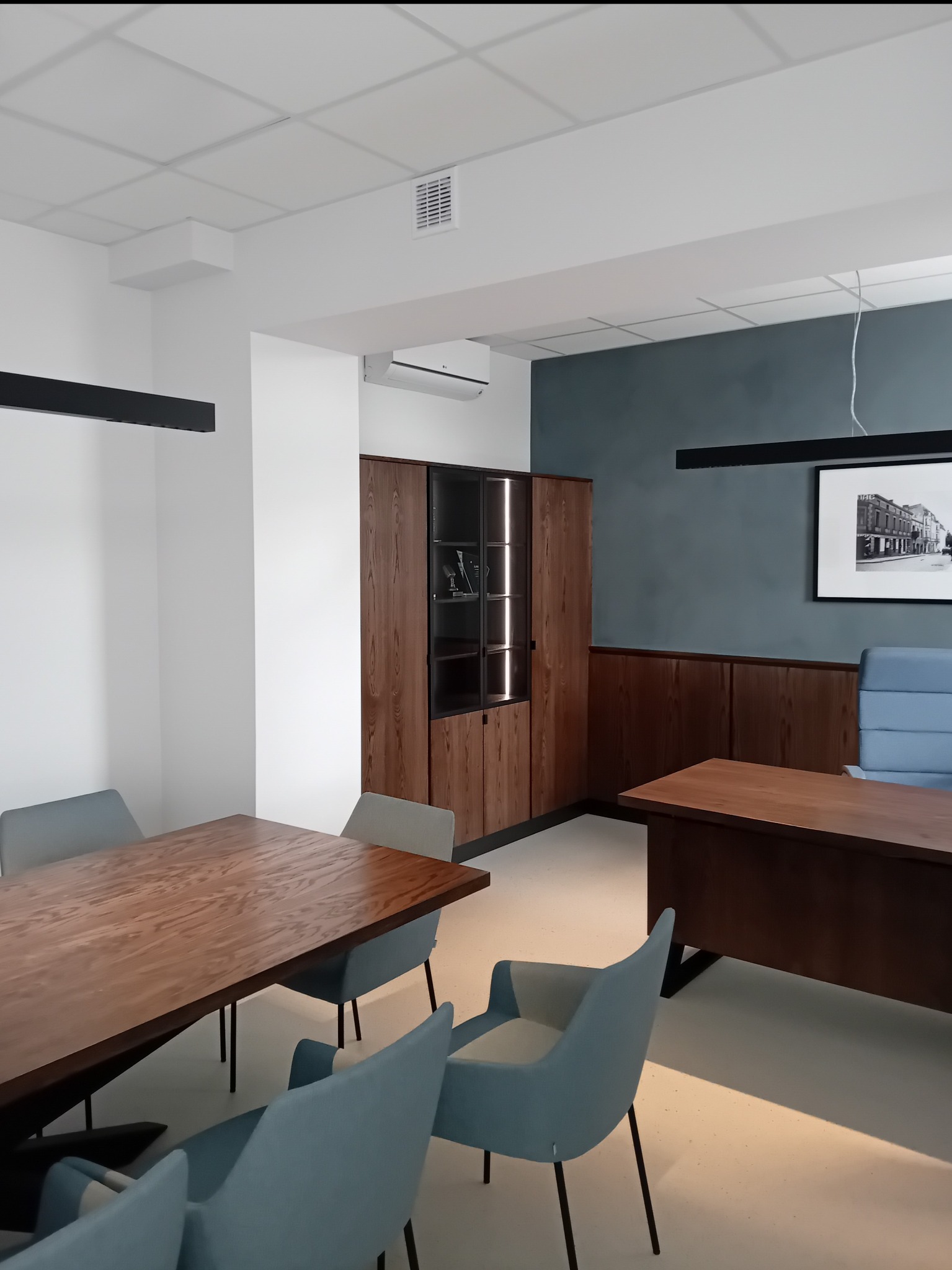Nowoczesny gabinet z drewnianym biurkiem i stołem konferencyjnym, niebieskie krzesła, ciemna ściana z obrazem i podświetlana witryna.