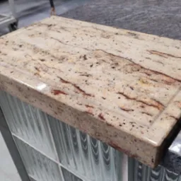 Beżowy blat z konglomeratu z brązowymi żyłkami, z wyfrezowanym rantem, zamontowany na konstrukcji z luksferów, widok z góry pod kątem.