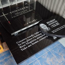 Kamieniarstwo GAP - Czarne, polerowane nagrobki z granitu: jeden w kształcie serca z wygrawerowanym portretem i niemieckim tekstem, drugi prostokątny z krzyżem i napisem 'Ave Maria', ustawione w warsztacie na tle...