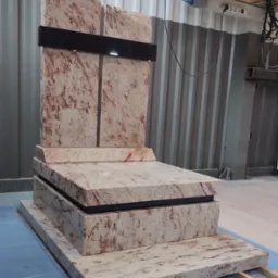 Nowoczesny pomnik z jasnego granitu z ciemnymi akcentami, ustawiony na drewnianych legarach w hali produkcyjnej.