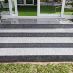 Zewnętrzne schody z szarego kamienia prowadzące do domu, z roślinami w donicach po bokach. Widoczne cztery stopnie i fragment trawnika.