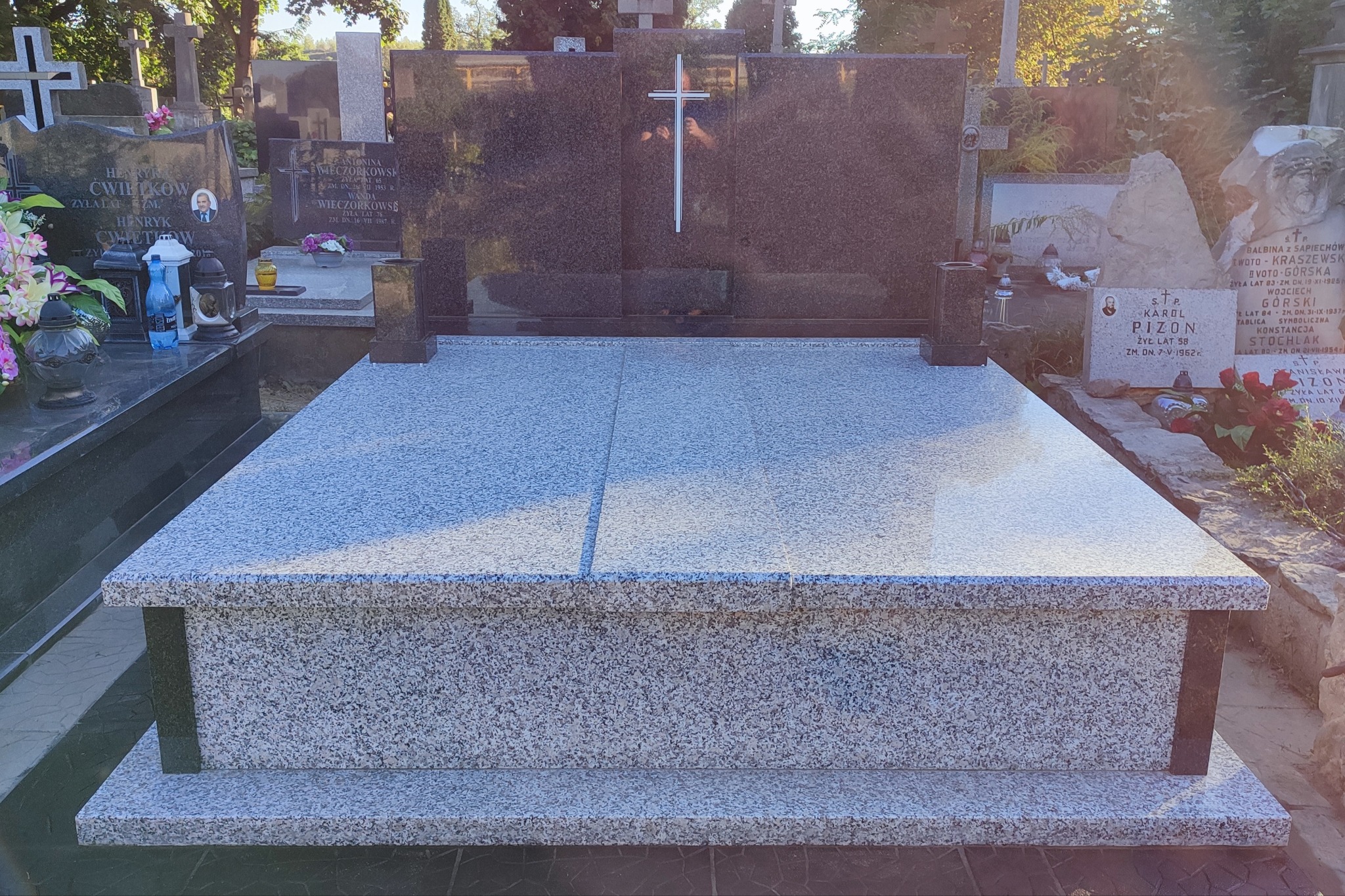 Granitowy pomnik nagrobny z krzyżem, widok z przodu, na cmentarzu w słoneczny dzień.