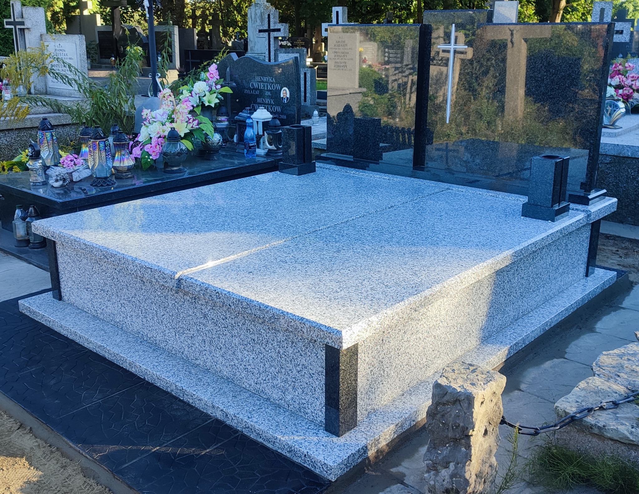 Jasnoszary, dwupoziomowy nagrobek z granitu z czarnymi elementami dekoracyjnymi, ustawiony na czarnej, wzorzystej płycie w otoczeniu innych nagrobków i zieleni cmentarnej.