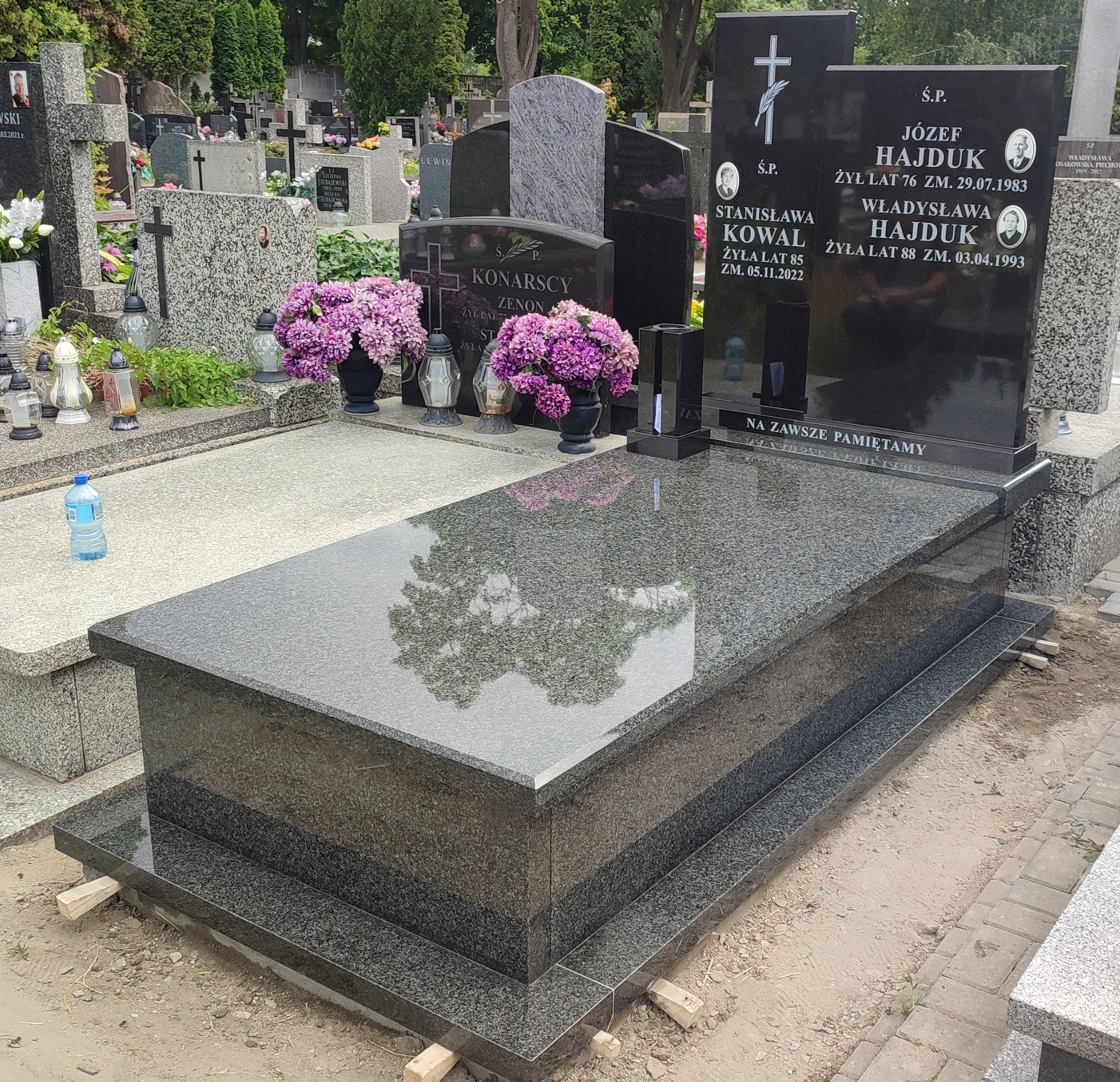 Ciemnoszary, wypolerowany nagrobek z dwoma pionowymi płytami z wygrawerowanymi napisami i zdjęciami, udekorowany fioletowymi kwiatami w czarnych wazonach, na tle innych nagrobków i zieleni cmentarza.