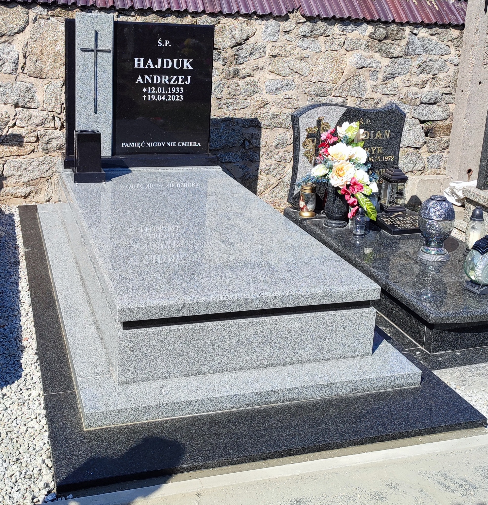 Granitowy nagrobek z czarną tablicą inskrypcyjną i krzyżem, z widocznymi datami urodzin i śmierci oraz napisem 'Pamięć nigdy nie umiera', na tle kamiennego muru.