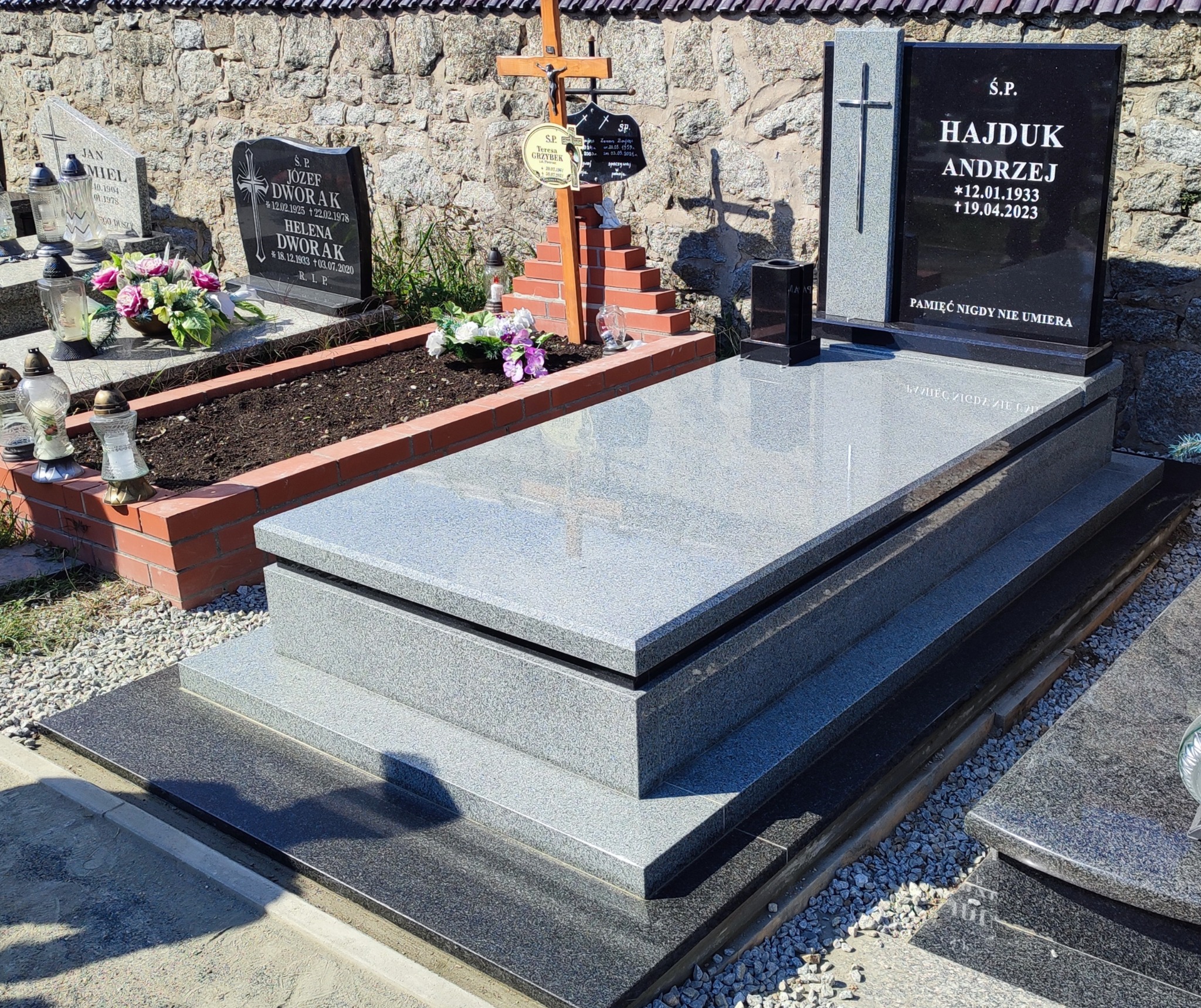 Nowoczesny, dwupoziomowy pomnik z szarego i czarnego granitu, z wygrawerowanym krzyżem i napisem, otoczony kamiennym murem i innymi nagrobkami na cmentarzu.