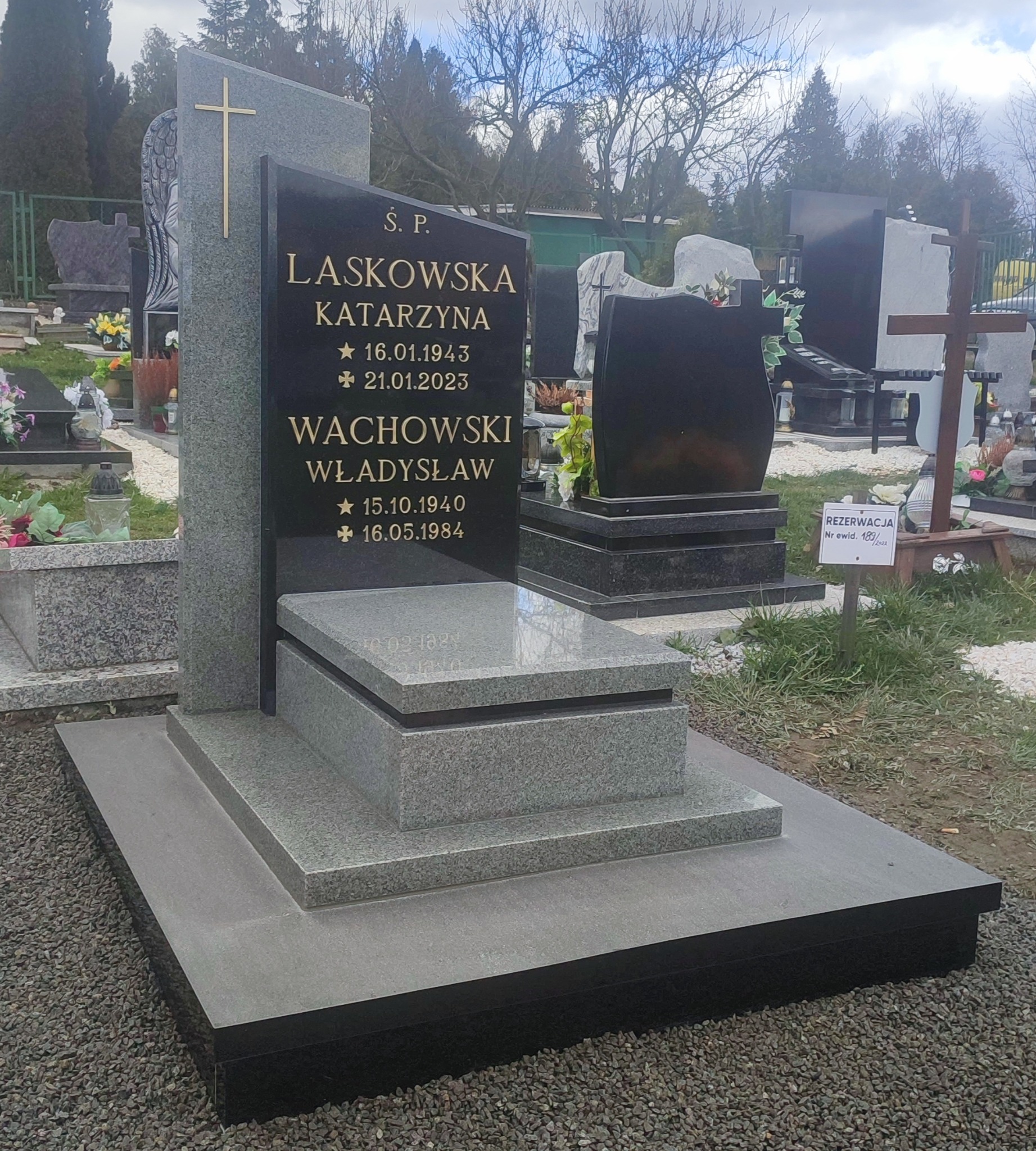 Nowoczesny, dwukolorowy pomnik z jasnoszarego i czarnego kamienia na cmentarzu, z wygrawerowanymi złotymi literami i krzyżem, otoczony drobnym żwirem.