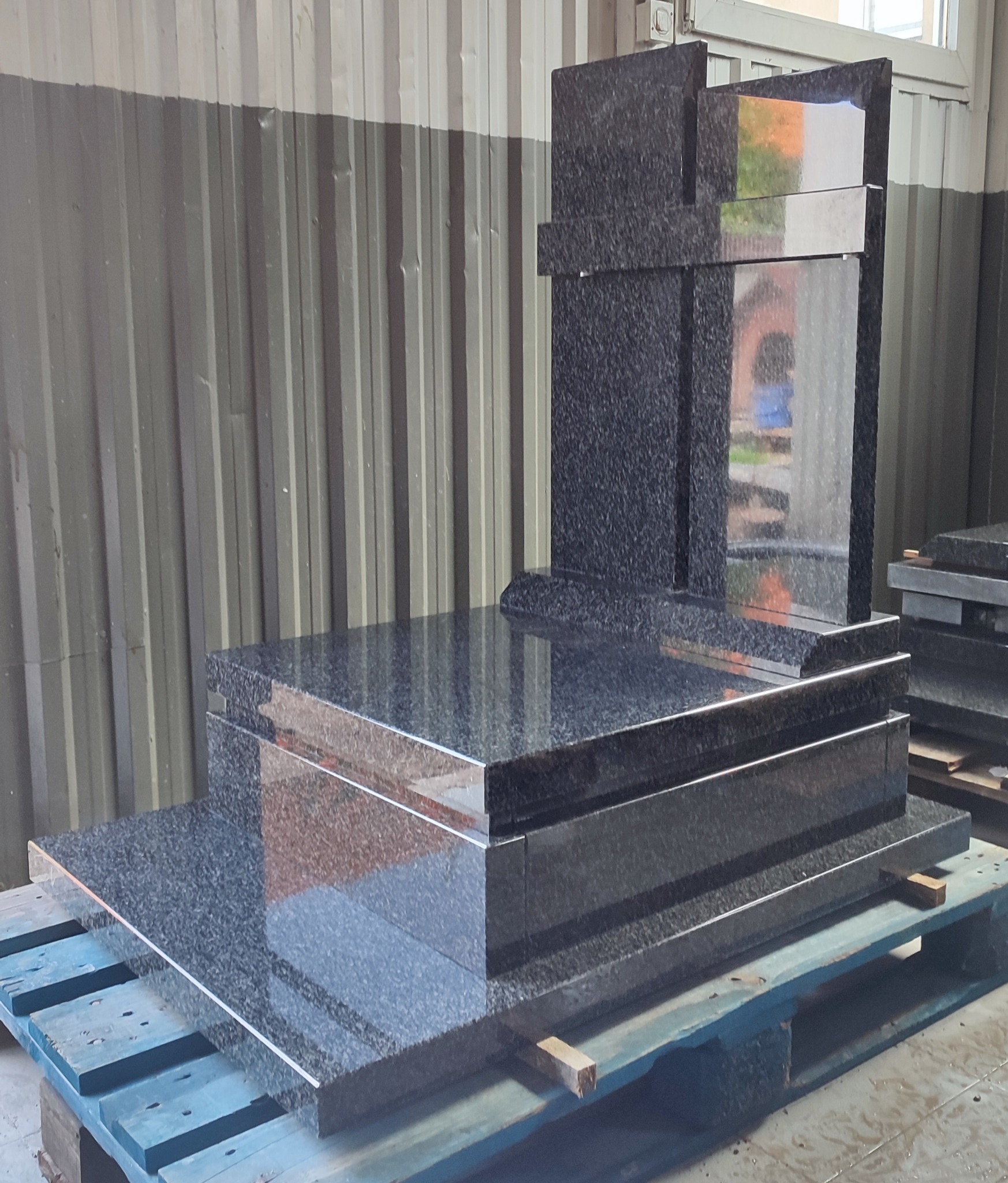 Czarny, polerowany nagrobek z granitu z krzyżem, na palecie w hali produkcyjnej, widoczne odbicia w kamieniu.