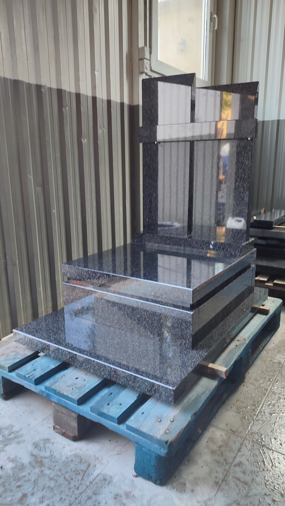 Czarny, granitowy pomnik na niebieskiej palecie w magazynie, widoczne odbicia światła na polerowanej powierzchni.