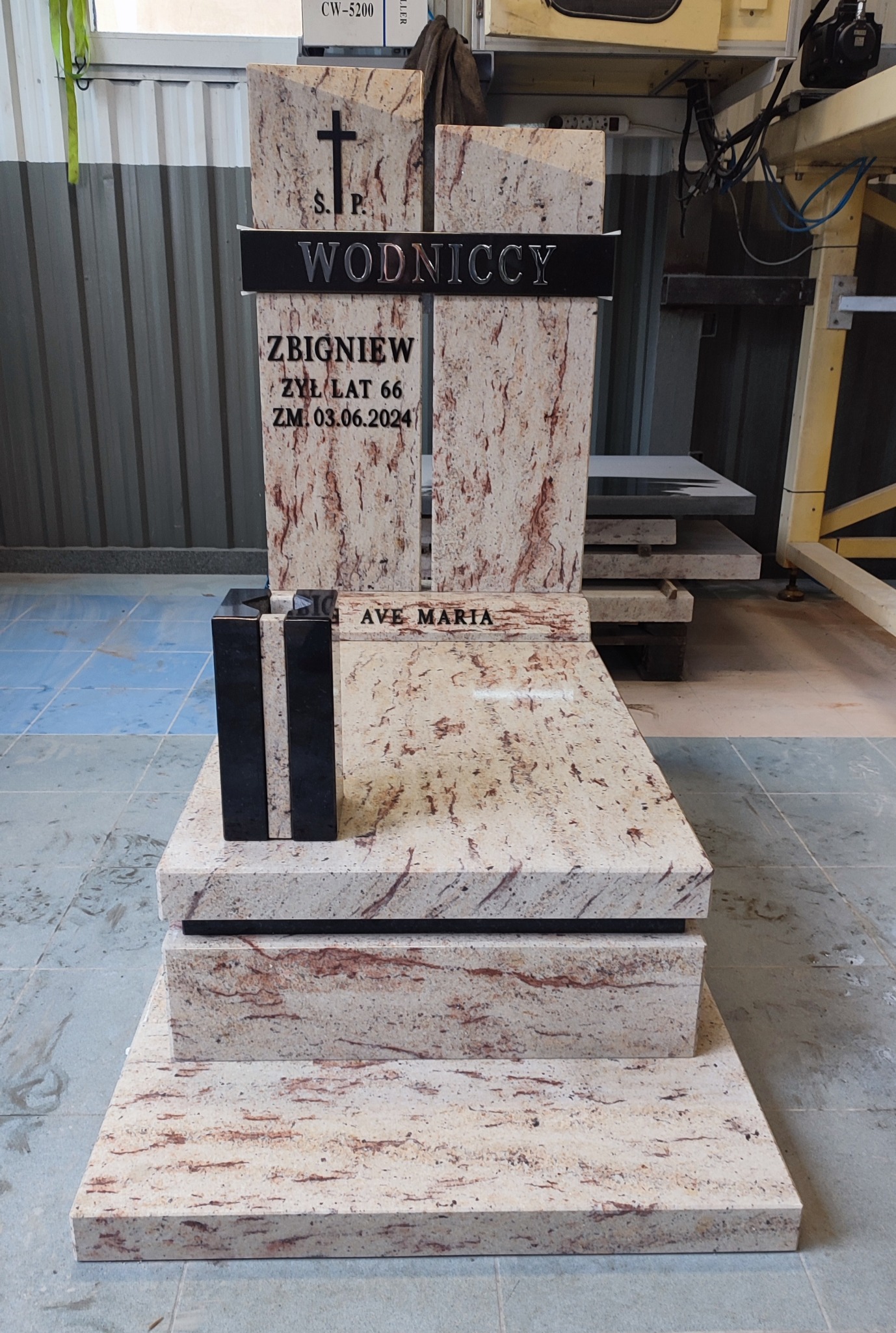 Jasny, granitowy pomnik z czarnymi elementami, z wygrawerowanym nazwiskiem 'Wodniccy' i datą śmierci 03.06.2024, ustawiony w warsztacie kamieniarskim, widoczne maszyny i płyty kamienne w tle.