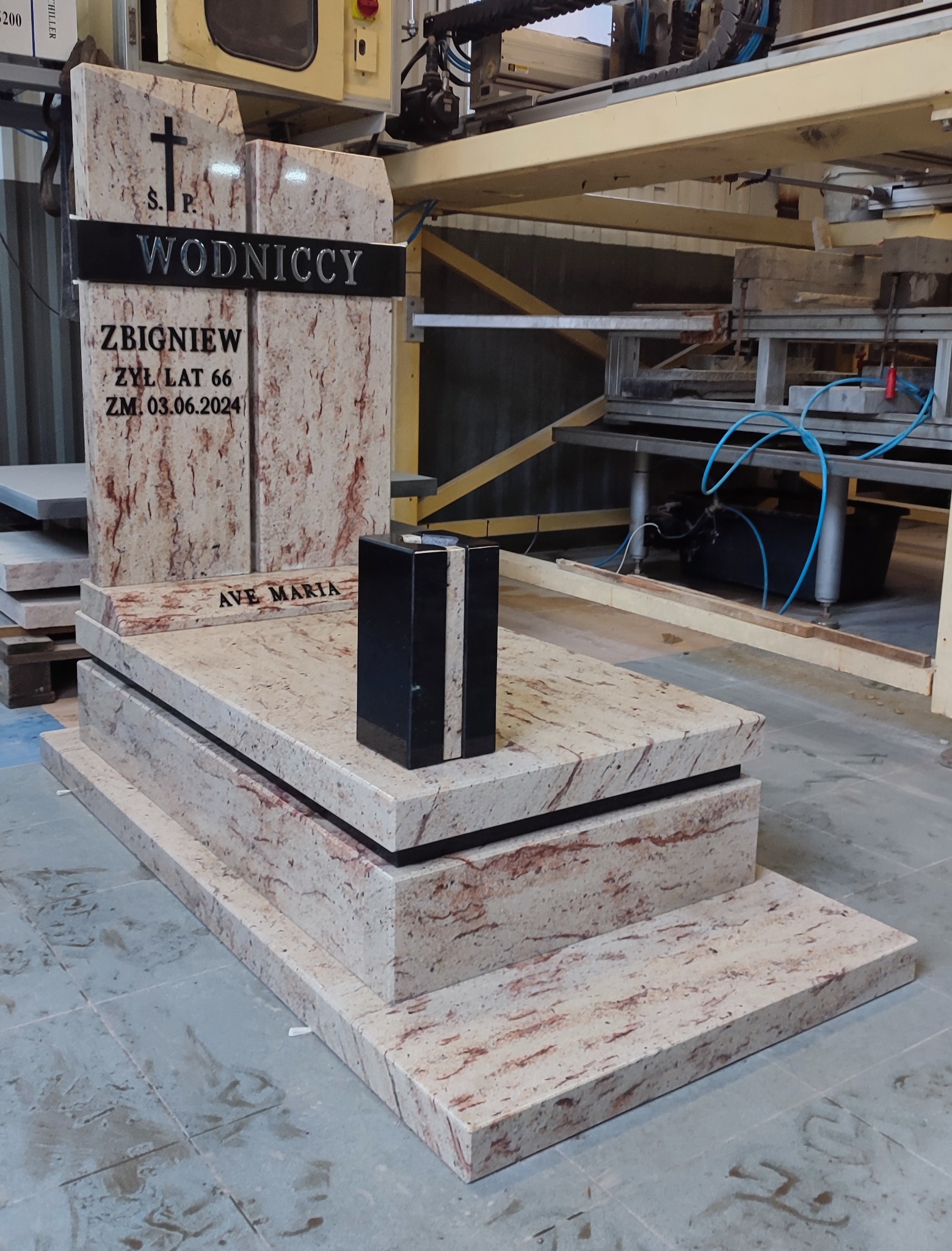 Jasny, granitowy pomnik z czarnym wazonem, widoczny w warsztacie kamieniarskim, z maszyną CNC w tle.