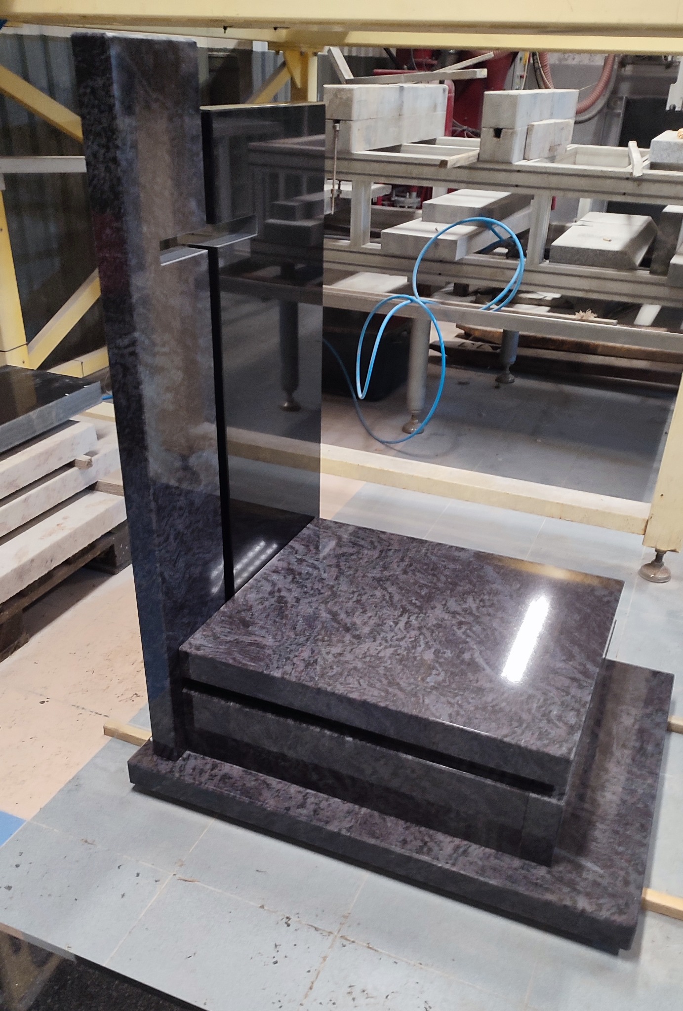 Surowy, ciemny nagrobek z granitu w trakcie produkcji, widoczny na tle hali produkcyjnej z elementami maszyn i surowych płyt kamiennych.