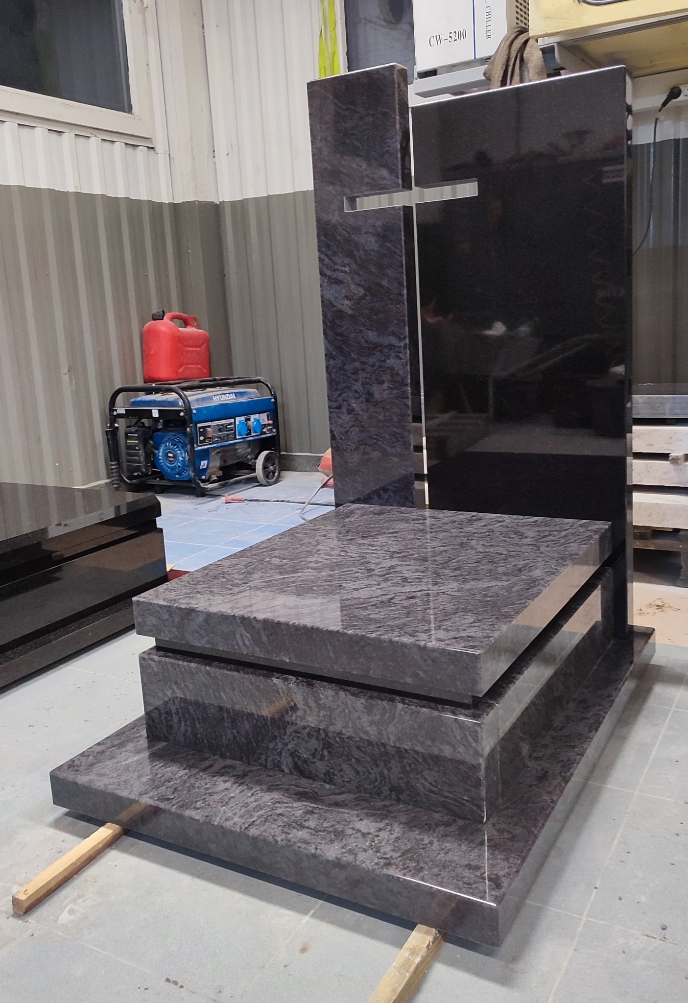 Wieloelementowy, granitowy pomnik z wyciętym krzyżem, ustawiony w warsztacie, widoczny agregat prądotwórczy i kanister.