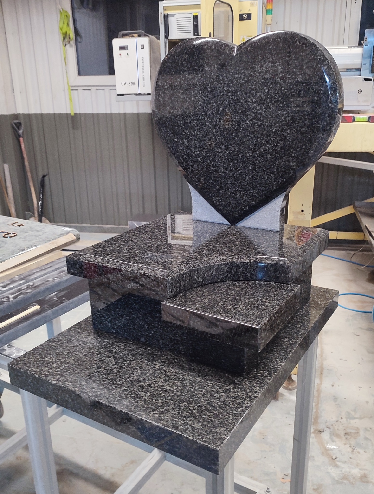 Czarny, granitowy nagrobek w kształcie serca, częściowo złożony w warsztacie, z widocznymi elementami konstrukcyjnymi i sprzętem do obróbki kamienia w tle.