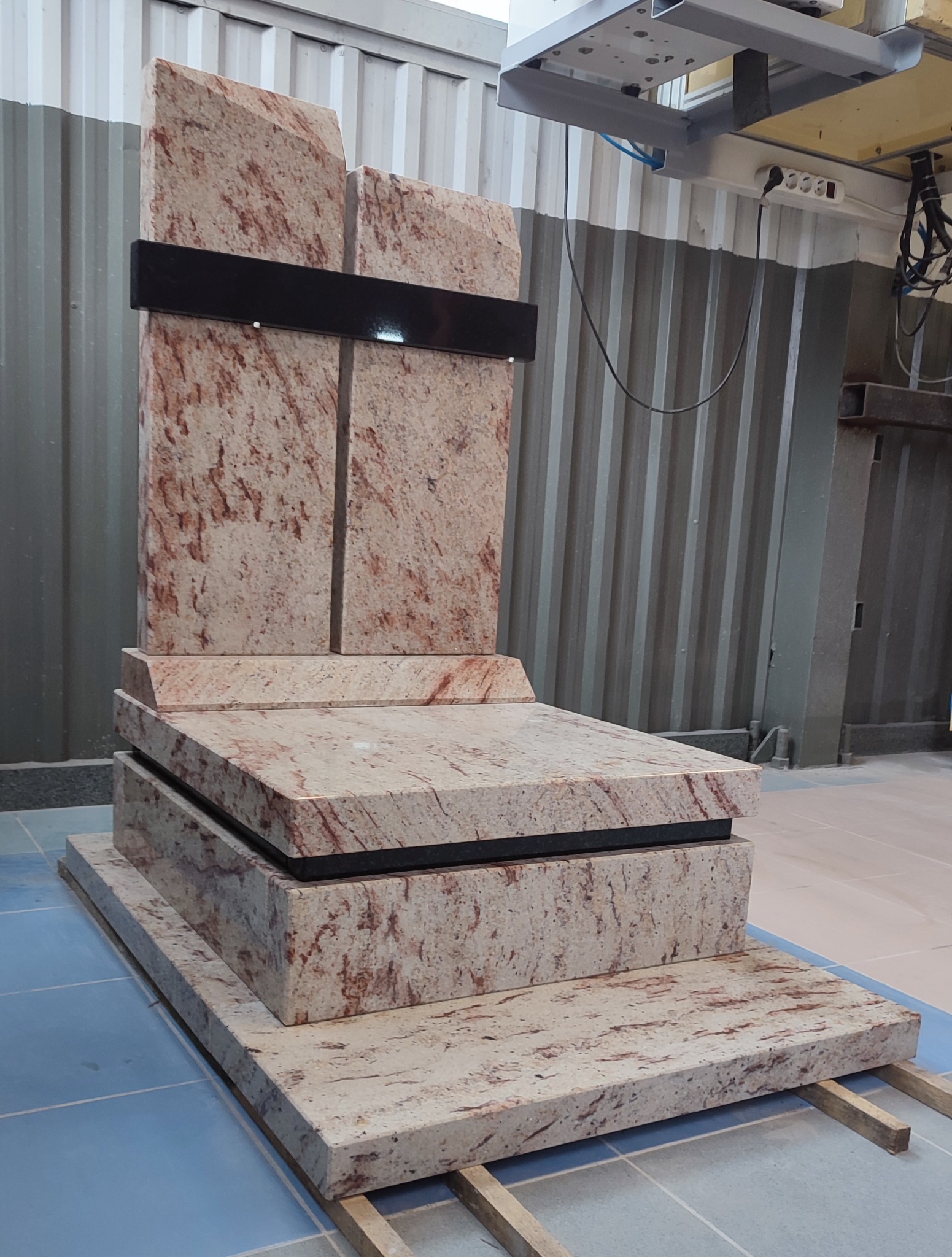 Nowoczesny pomnik z jasnego granitu z ciemnymi akcentami, ustawiony na drewnianych legarach w hali produkcyjnej.